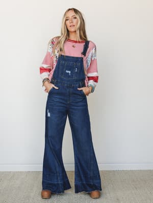 Wide-leg dark wash denim overalls styled over a floral-and-stripe layered long sleeve top, accessorized with turquoise and beaded bracelets and brown clogs — the kind of effortless, lived-in outfit that feels equally at home at a Saturday market or a