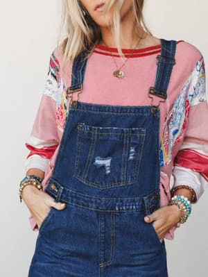 A relaxed, lived-in look built around dark wash denim overalls layered over a red striped long-sleeve top with patchwork sleeves — styled with stacked bracelets and a delicate gold necklace for that effortlessly collected boho feel.