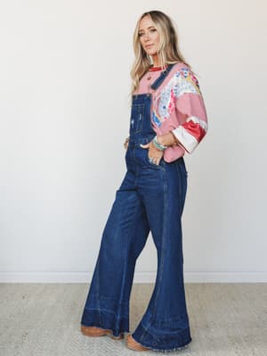 Wide-leg denim overalls in a deep indigo wash styled over a mixed-print graphic tee, paired with turquoise beaded bracelets and clogs — effortlessly vintage with a free-spirited layered look.