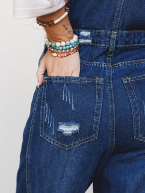 A close-up detail shot of dark wash distressed denim overalls, hand tucked in back pocket and stacked boho bracelets stealing just as much attention as the denim itself.