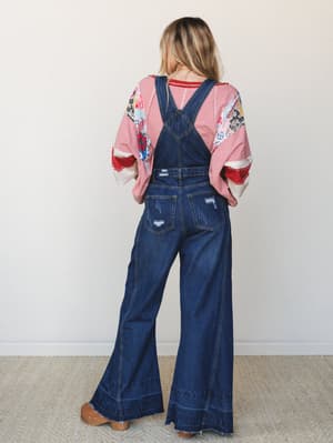 Shot from the back, a model wears wide-leg dark wash denim overalls over a patchwork graphic top with red stripe layering — the kind of effortlessly collected outfit that somehow looks like it took zero effort and total intention at the same time.