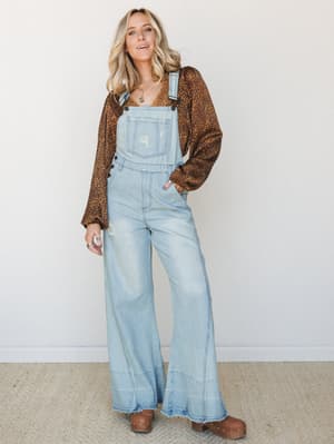 Wide-leg light wash denim overalls styled over a long-sleeve leopard print top, finished with brown clogs and layered necklaces — a lived-in, throwback-cool look that feels like a weekend with nowhere specific to be and everywhere to go.