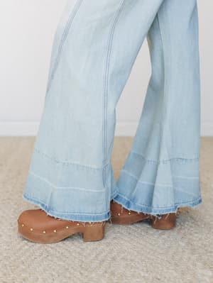 A close-up of wide-leg bell bottom jeans in light wash denim with a frayed hem, paired with studded tan leather clogs — very much the vibe of a '70s record collection brought back to life.