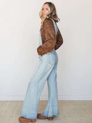 A model shot in three-quarter profile, glancing back over her shoulder — light wash wide-leg denim overalls paired with a rust leopard print top and brown clogs give this look an easy 70s-meets-everyday energy.