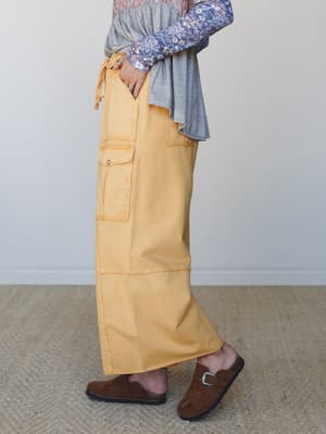 A side-profile shot of a marigold yellow wide-leg cargo pant styled with a layered gray top and floral-sleeved underlayer, finished with brown suede clogs on a jute rug — effortlessly relaxed and a little earthy.