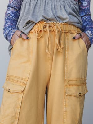 A close-up detail shot of wide-leg cargo pants in a washed mustard yellow, styled with a layered grey tee and floral print top — hands in pockets, relaxed and completely lived-in.