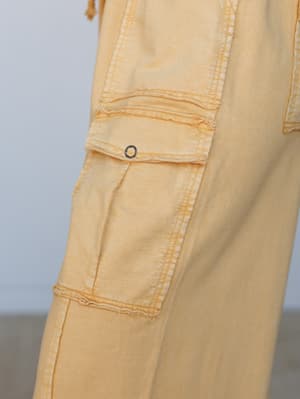 A close-up detail shot of washed mustard cargo pants showing the side pocket construction — raw-edge seams, an eyelet ring closure, and that lived-in, sun-faded texture that makes them look like they've already been somewhere interesting.