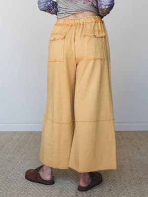 A back-view shot of wide-leg cargo pants in washed mustard, styled with a floral-sleeve top and brown leather clogs — relaxed and expressive with an easy, lived-in feel.