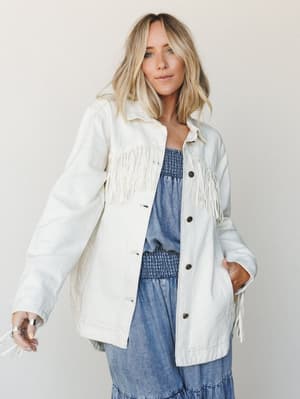A cream fringe jacket layered over a blue smocked midi dress — effortlessly western-boho, shot against a soft neutral wall with a relaxed, confident energy.