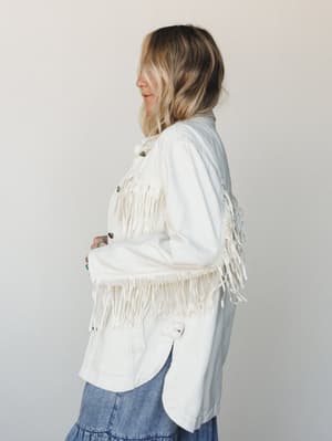 A side-profile shot of a model in a white denim fringe jacket layered over a tiered blue denim skirt — the kind of outfit that feels effortlessly western without trying too hard.
