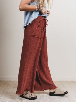 Woman wearing rust-colored wide-leg pants with a light blue top and sandals on a neutral background
