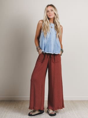 Woman wearing a light blue sleeveless top and rust-colored wide-leg pants against a plain background