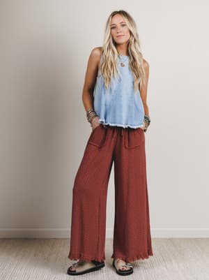 Woman wearing a light blue sleeveless top and rust-colored wide-leg pants against a plain background