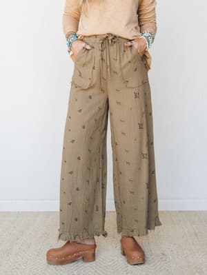 Wide-leg floral pants in an earthy olive tone styled with a soft peach long-sleeve top, stacked turquoise bracelets, and brown clogs — the kind of outfit that looks effortless but clearly wasn't an accident.