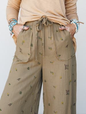 A close-up of wide-leg pants in a warm olive-tan fabric scattered with tiny botanical floral sprigs, styled with a sandy long-sleeve top and layers of turquoise and pearl bracelets — the kind of outfit that feels like a slow morning done right.