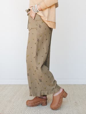A side-profile shot of wide-leg floral pants in an earthy olive tone, styled with a peach oversized top, stacked bead bracelets, and warm brown clogs — the kind of effortless, put-together look that feels intentional without trying too hard.