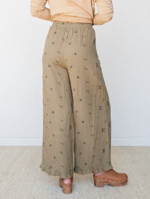 A back view of wide-leg pants in a warm khaki-tan with a scattered ditsy floral print, styled with a peach long-sleeve top and brown wooden clogs — the kind of easy, earthy outfit that feels like a farmers market morning.