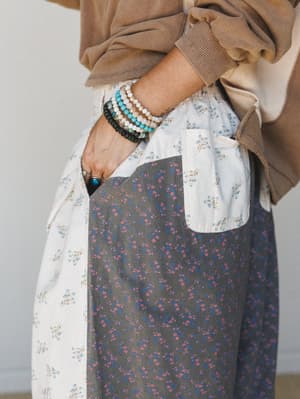 A close-up detail shot of mixed floral print wide-leg pants — two contrasting ditsy floral panels in cream and charcoal — styled casually with a washed tan sweatshirt and layered bead bracelets with a turquoise ring.