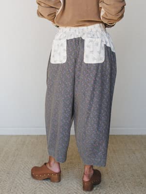 A back-view shot of wide-leg cropped pants in a dusty olive floral print with contrast cream floral patch pockets at the waist, styled with a camel sweatshirt and wooden clogs — effortlessly relaxed with a collected, vintage charm.
