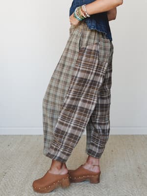 A side-profile shot of relaxed plaid pants in earthy brown and sage tones, styled with a denim crop top, stacked bead bracelets, and warm tan clogs — the kind of outfit that feels vintage-thrifted but completely intentional.