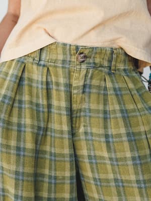 A close-up detail shot of high-waisted plaid wide-leg pants in a faded olive and slate blue plaid, styled with a cream linen tee and a peek of beaded jewelry — earthy, relaxed, and the kind of pants you reach for on a good day.