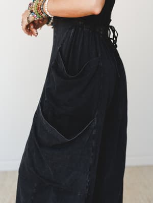 A close-up detail shot of a washed black cargo jumpsuit, highlighting the oversized curved patch pockets and elastic waistband — styled with a stack of colorful beaded bracelets and rings that give it an effortless boho personality.