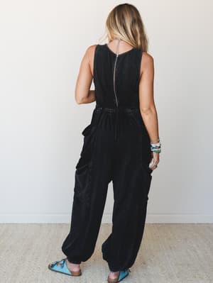 Shot from the back, a model wears a washed black cargo jumpsuit with wide, relaxed legs and patch pockets — the kind of effortless one-and-done piece that looks equally at home at a weekend market or a casual night out, styled with turquoise sandals
