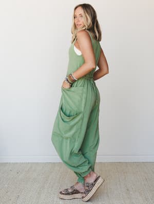 A relaxed, lived-in sage green cargo jumpsuit styled from the side with platform leopard sandals and a stack of beaded bracelets — effortlessly boho, the kind of outfit that looks like you tried just enough.