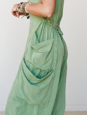A close-up side shot of a sage green washed jumpsuit, styled with stacked beaded bracelets — the focal point is an oversized cargo pocket that drapes with a soft, lived-in ease.