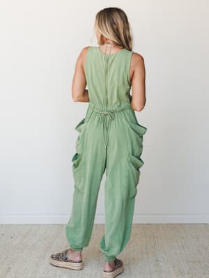 A sage green washed cargo jumpsuit shot from the back, with roomy patch pockets and a drawstring waist — the kind of one-and-done outfit that looks effortless but clearly made a decision.