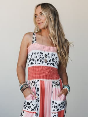 A smocked, sleeveless jumpsuit in a coral and white color-block with bold leopard print paneling — layered with stacked boho bracelets and a delicate pendant necklace for an effortlessly expressive summer look.