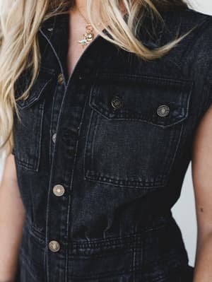 A close-up torso shot of a black washed denim jumpsuit with antique brass snap buttons and a chest pocket, styled with a delicate gold star charm necklace — the kind of piece that looks equally at home on a Saturday farmer's market run or a night out