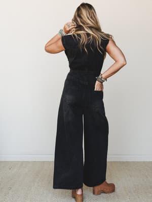 Shot from behind, a model wears a black washed wide-leg denim jumpsuit styled with stacked turquoise bracelets and tan studded clogs — the kind of effortless, put-together look that works as hard as it plays.
