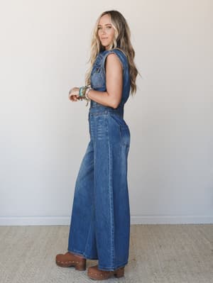 A model shot in profile wearing a medium wash sleeveless denim jumpsuit with wide-leg trousers, styled with stacked beaded bracelets and wood-studded clogs — easy, confident, and just the right amount of vintage cool.