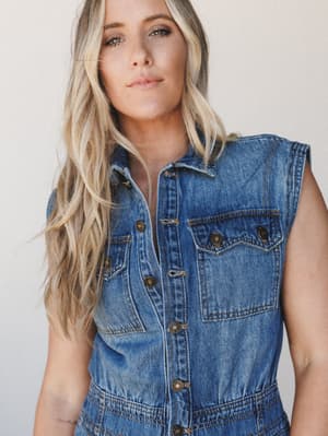 A medium wash denim jumpsuit worn by a blonde model against a soft white studio backdrop — effortlessly cool with a lived-in, vintage feel that reads equal parts casual and intentional.