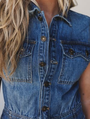A close-up detail shot of a medium wash denim jumpsuit with vintage-style snap buttons and chest pockets — the kind of piece that looks like it's been living in someone's closet in the best possible way.