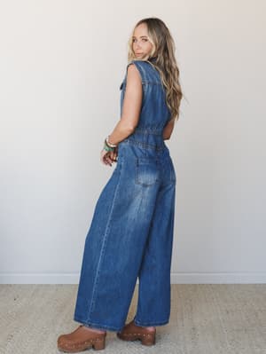 A wide-leg denim jumpsuit in medium wash photographed from a three-quarter back angle — relaxed and a little retro, styled with brown clogs and a stack of bracelets that feel completely lived-in.