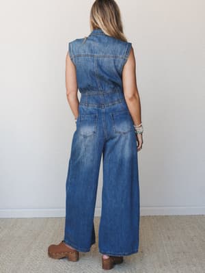 A wide-leg sleeveless denim jumpsuit shot from the back, styled with brown clogs and stacked bracelets — effortless and a little retro, like something you'd grab off the hanger and never want to take off.