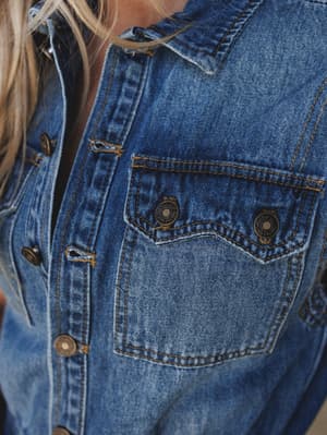A close-up detail shot of a medium wash denim jacket or jumpsuit, showing the classic chest pocket, contrast topstitching, and worn-in texture — the kind of piece that looks better the more you live in it.