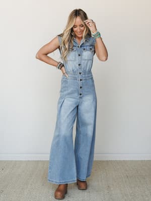 A wide-leg denim jumpsuit in light blue wash styled with brown clogs and stacked beaded bracelets — the kind of outfit that does all the work without trying too hard.