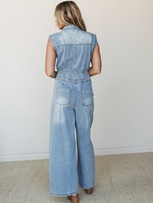 A back-view shot of a light-wash denim jumpsuit with wide-leg pants, a cinched elastic waist, and a sleeveless silhouette — easy and a little retro, styled with brown heeled sandals against a clean studio backdrop.