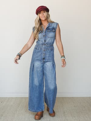 A wide-leg denim jumpsuit styled with a burgundy newsboy cap, stacked beaded bracelets, and brown clogs — the kind of effortlessly put-together look that makes a Saturday errand run feel intentional.