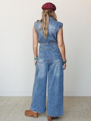 A back-view shot of a wide-leg denim jumpsuit styled with a burgundy beret, stacked boho bracelets, and tan clogs — the kind of outfit that feels effortless but clearly thought about.