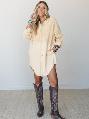 An oversized corduroy tunic dress in a soft washed butter yellow is styled with tall dark brown cowboy boots and a stack of colorful beaded bracelets — the kind of effortless outfit that somehow looks like you planned it.