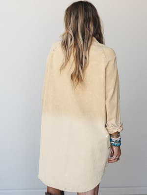 Shot from the back, a corduroy tunic dress in a soft mustard dip-dye wash hits mid-thigh with a lived-in, effortless feel — stacked turquoise bracelets and a silver ring are the only accessories she needs.