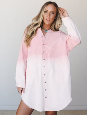 An oversized corduroy tunic dress in a soft pink dip-dye wash, styled casually with boho jewelry against a clean studio backdrop — effortless and a little dreamy.