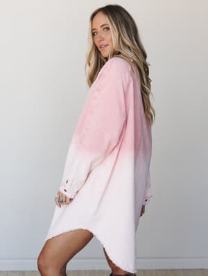 A breezy corduroy tunic dress in a soft pink dip-dye wash — worn oversized and effortless, styled with stacked rings and a glance back over the shoulder like she's already halfway out the door.