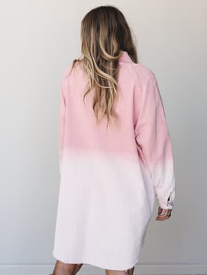 Shot from behind, a model wears an oversized corduroy tunic dress in a soft pink dip-dye wash — deeper rose at the top fading to barely-there blush at the hem, with a raw-edge finish that keeps it feeling relaxed and real.