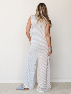 Shot from behind, a model wears a relaxed striped wide-leg jumpsuit in soft blue and beige, styled with stacked boho bracelets and suede clogs — the kind of effortless one-and-done outfit that looks like you tried just enough.