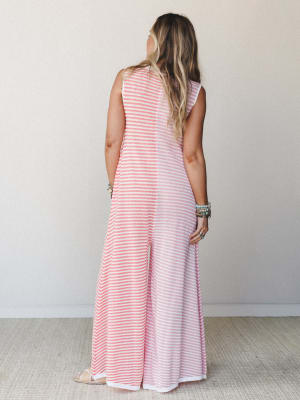 Shot from the back, a model in a floor-length pink and white striped sleeveless jumpsuit with wide legs stands against a soft neutral wall — the stacked turquoise and mixed-metal bracelets and strappy sandals giving it that effortless, collected summ